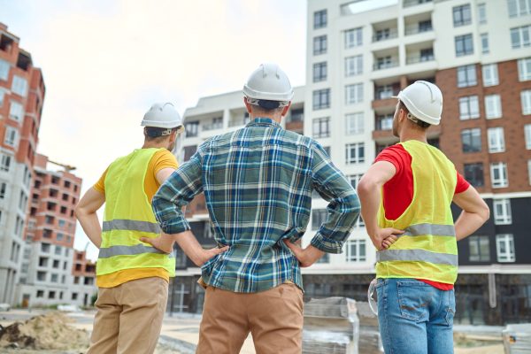 Result. Back view of three men in protective helmets holding hands on his waist standing at construction site looking at buildings under construction during day